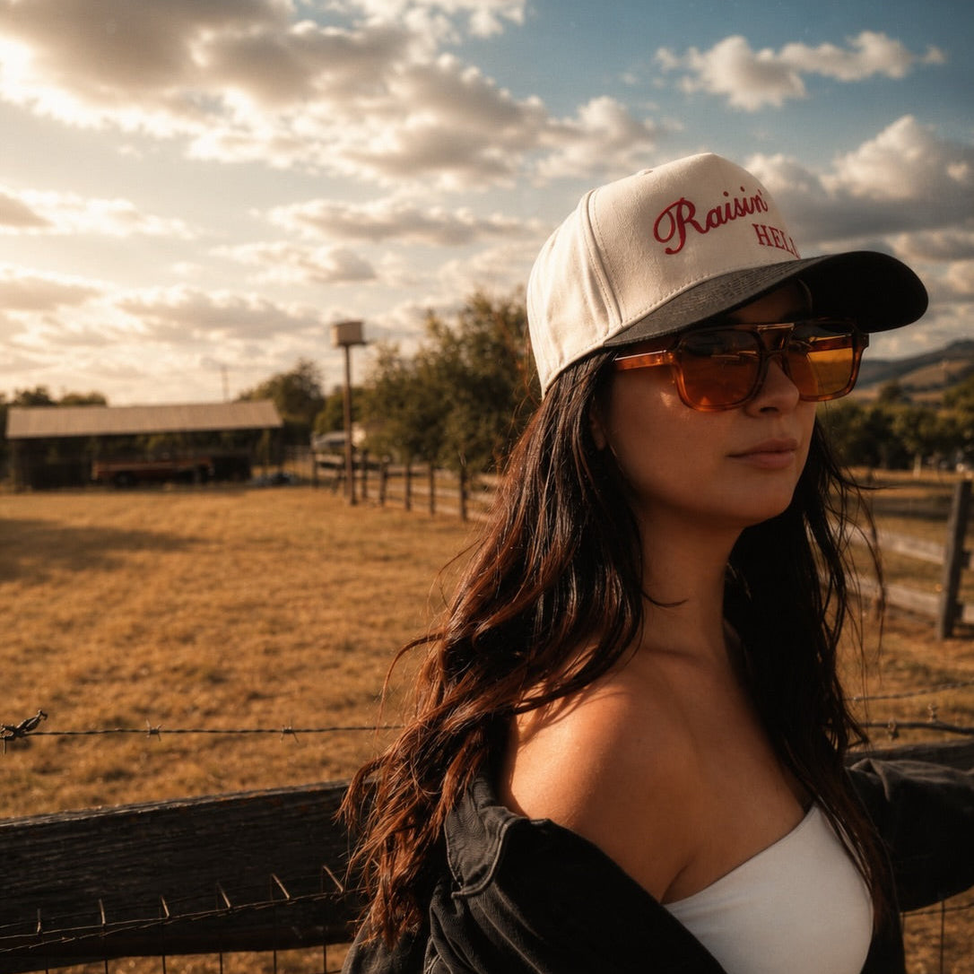 A woman in a white cap with red embroidered text, orange-tinted sunglasses, a white top, and a black denim jacket stands outdoors in a sunny field near a fence.